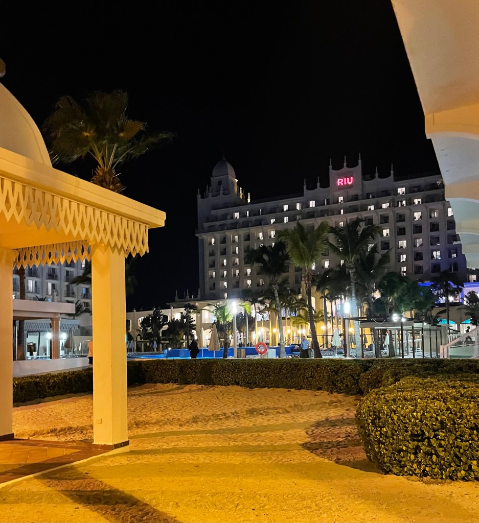 Nachtafbeelding van het Riu Palace Aruba resort met verlichte gebouwen en palm bomen, gezien vanuit de tuin.