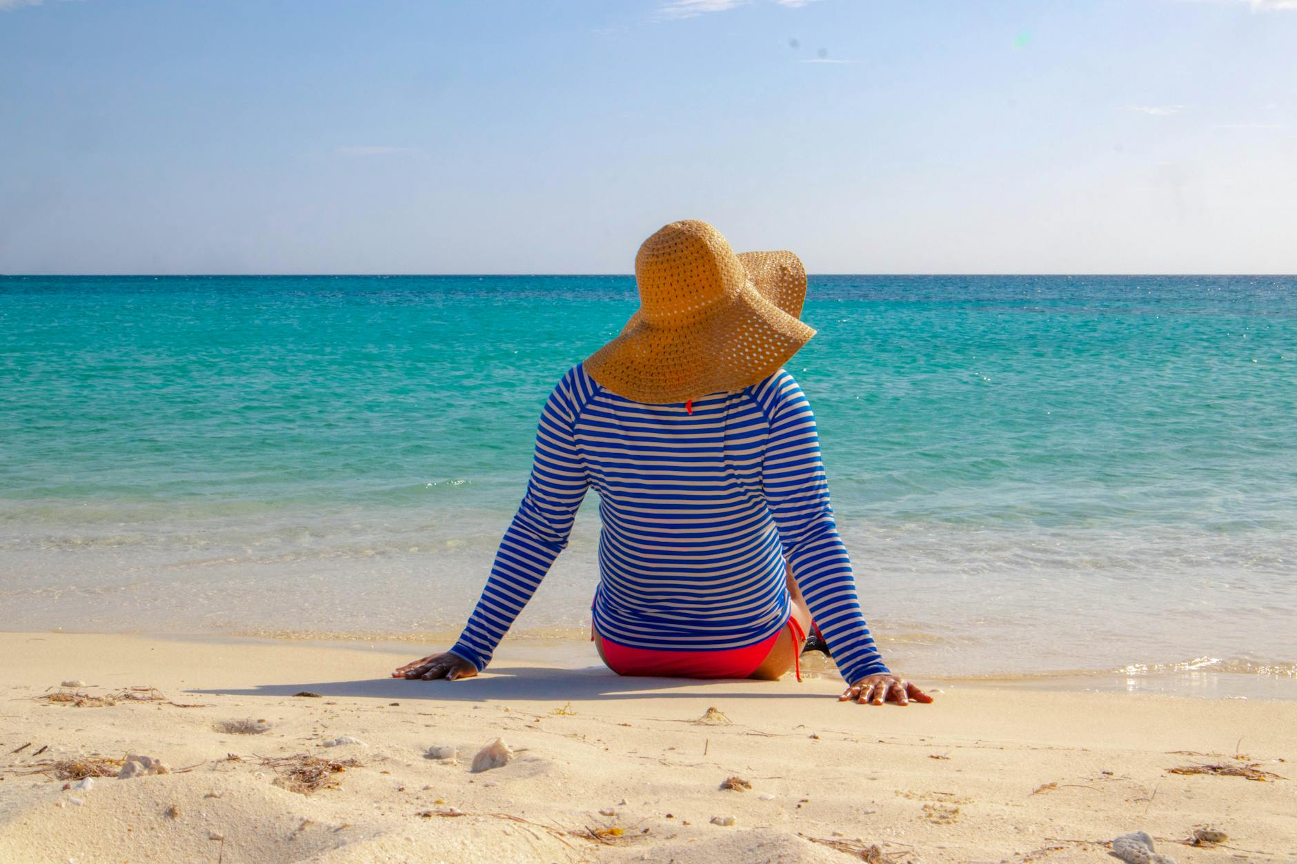Een persoon in een gestreept shirt en een grote strohoed zit op het strand en kijkt naar de oceaan onder een helderblauwe lucht.
